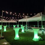 Outdoor nighttime event space with white canopy tents, string lights, and glowing green cocktail tables on the grass in front of a dark sky.