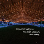 Large outdoor tent lit by warm string lights over a grassy field, set up for a concert tailgate at Mile High Stadium.