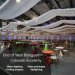 Event hall set for an End of Year Banquet at Colorado Academy with string lights, white fabric drapery, and colorful cocktail tables.