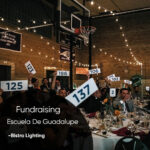 Audience at a charity fundraiser auction raising bid cards in a banquet hall with string lights and tables set for a meal, for Escuela De Guadalupe.